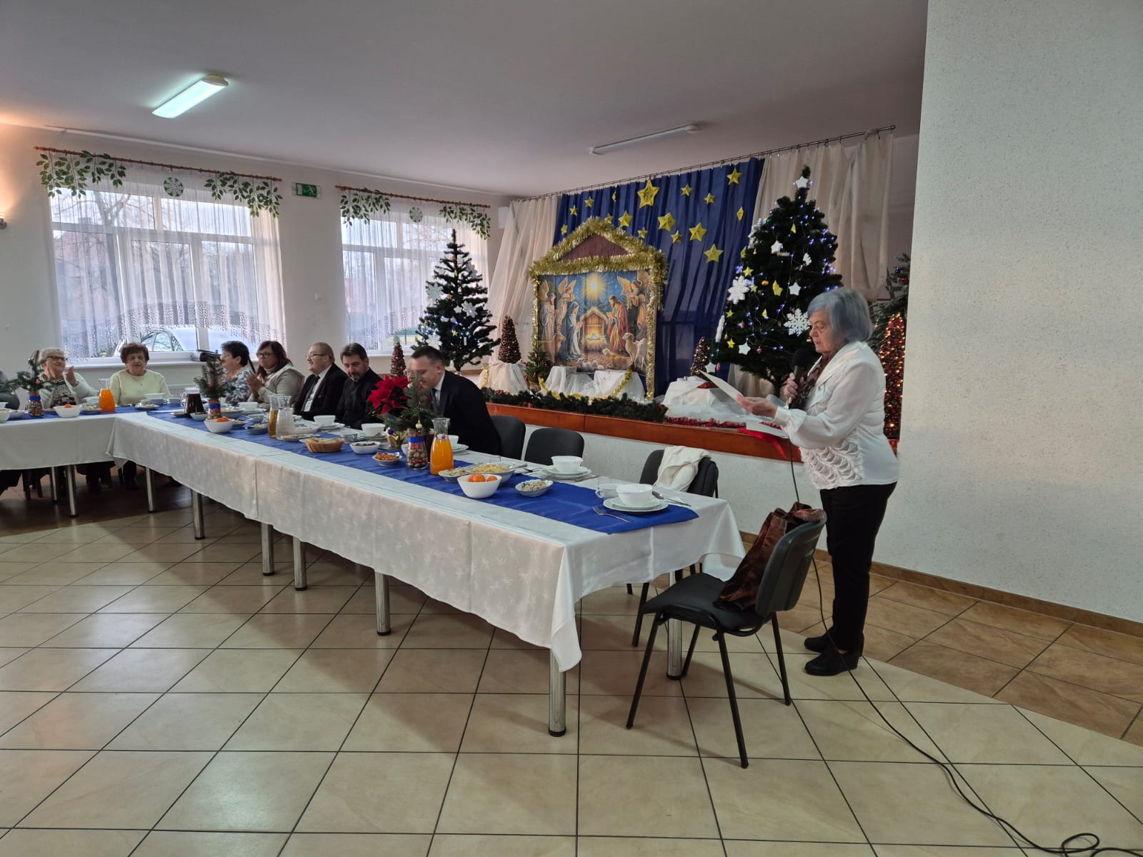 Przy uroczystym stole świątecznym, z&nbsp;tyłu scena ustrojona w&nbsp;zimowe choinki i&nbsp;żłobek