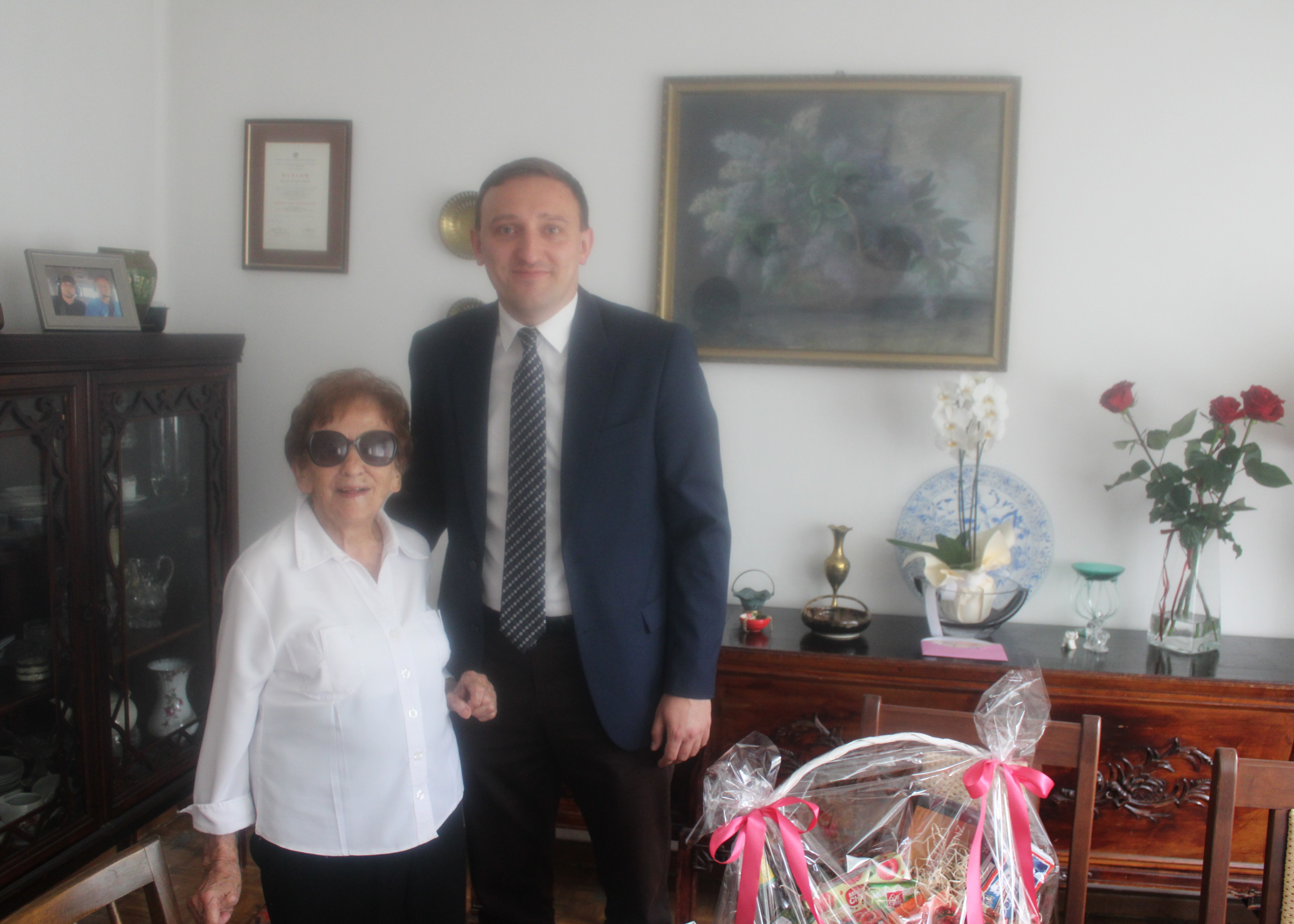 Kobieta w białej koszuli, obok pan w czarnym garniturze, na stole kosz prezentowy