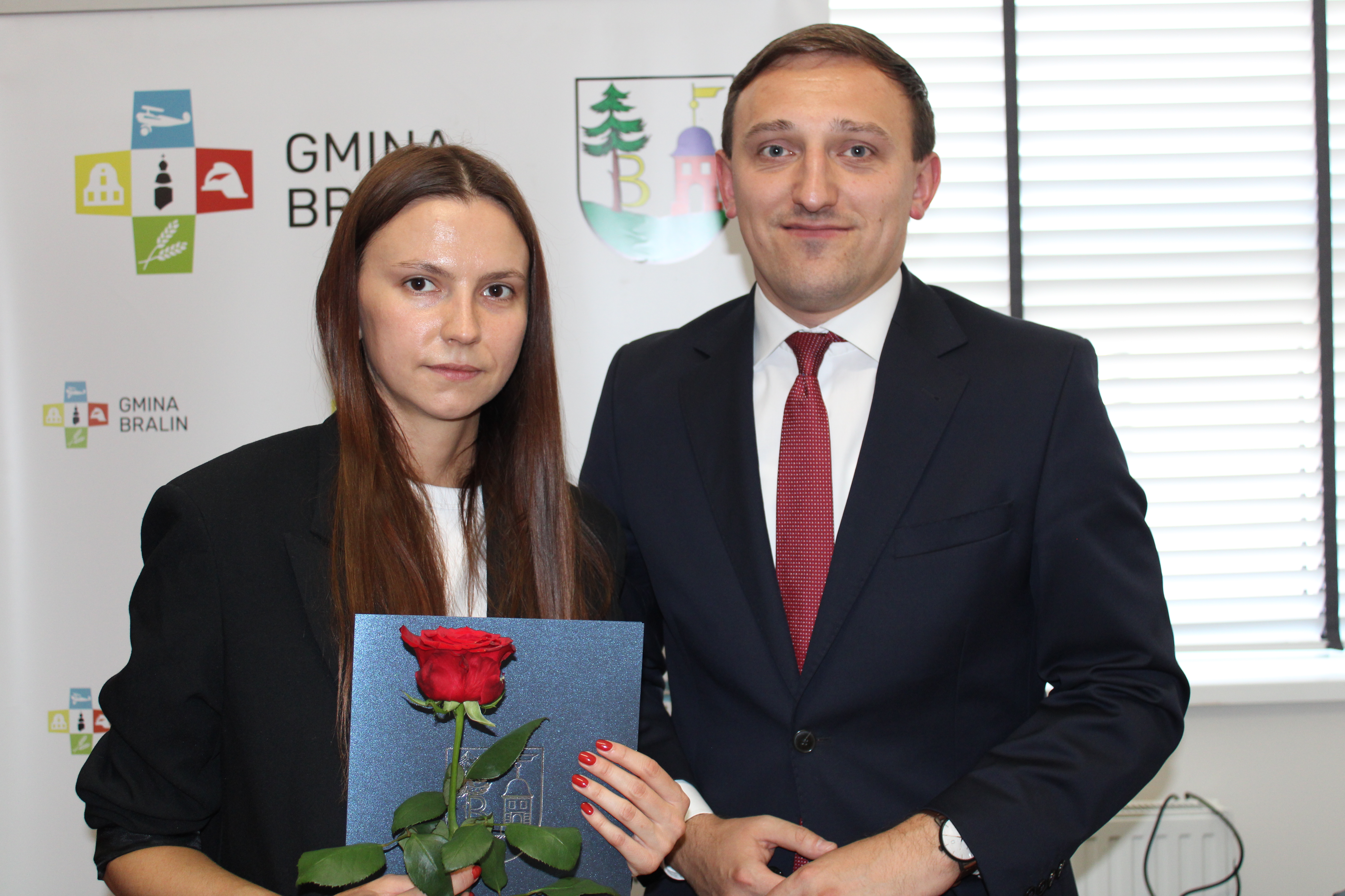 Kobieta w czarnej marynarce z różą w dłoni obok mężczyzna w garniturze