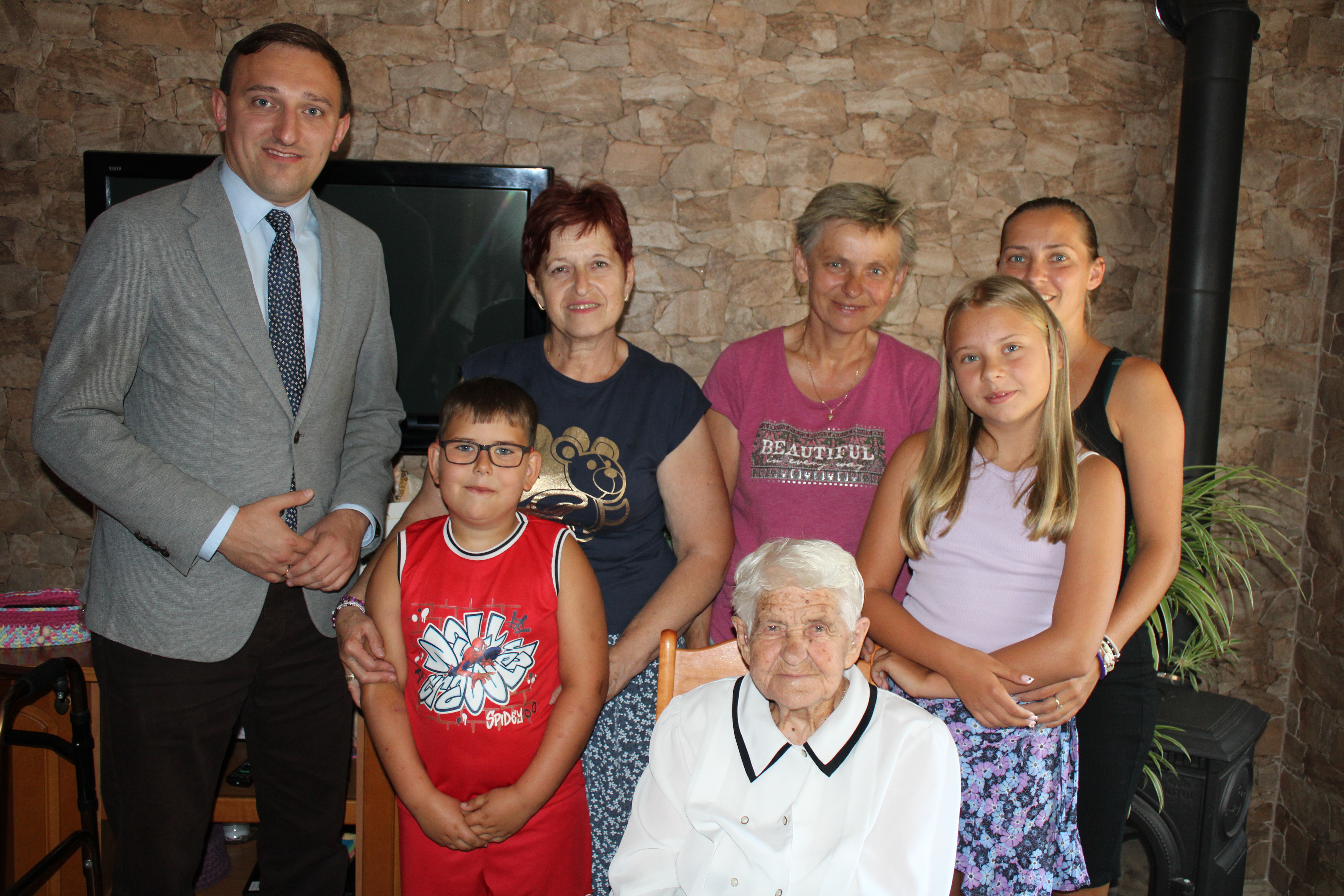 Mężczyzna w&nbsp;szarej marynarce, z&nbsp;prawej strony na&nbsp;środku starsza pani w&nbsp;białej koszuli, obok rodzina